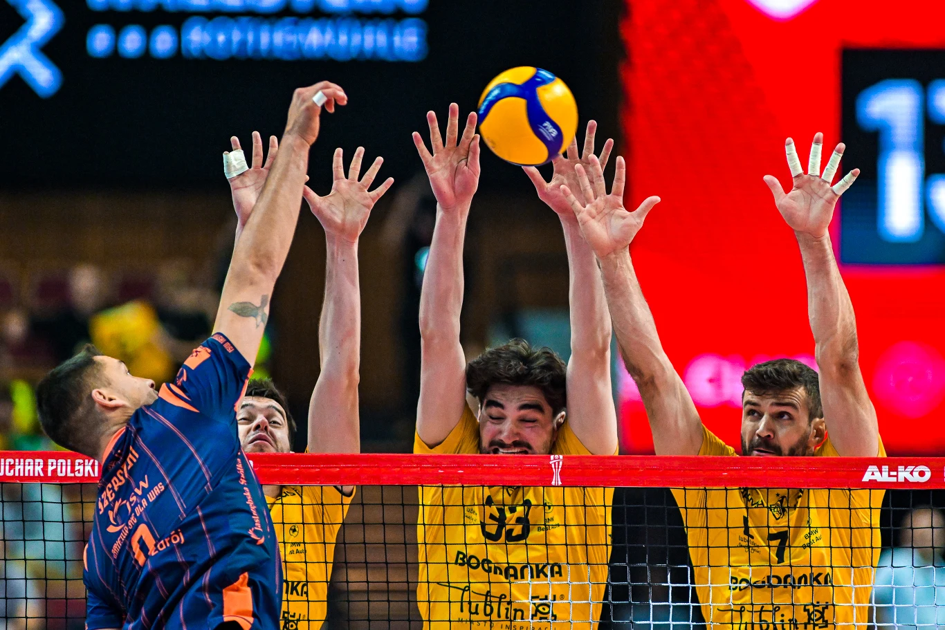 Fragment meczu siatkówki, w którym trzech zawodników drużyny ubranej na żółto próbuje zablokować atak siatkarza drużyny przeciwnej uderzającego piłkę nad siatką.