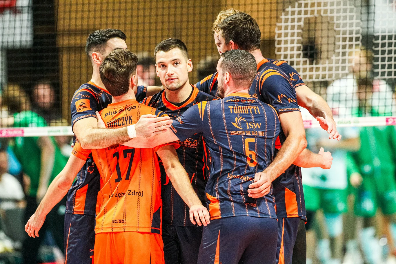 Nicolas Szerszeń (w środku) i siatkarze JSW Jastrzębskiego Węgla Grupa zawodników drużyny siatkarskiej w pomarańczowo-granatowych strojach celebruje zdobycie punktu, obejmując się nawzajem na boisku, w tle widzowie i siatka.