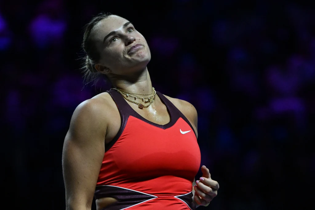 Kobieta w sportowym stroju tenisowym na ciemnym tle, pokazująca emocje niezadowolenia lub frustracji, stojąca z głową lekko uniesioną i spojrzeniem skierowanym do góry.