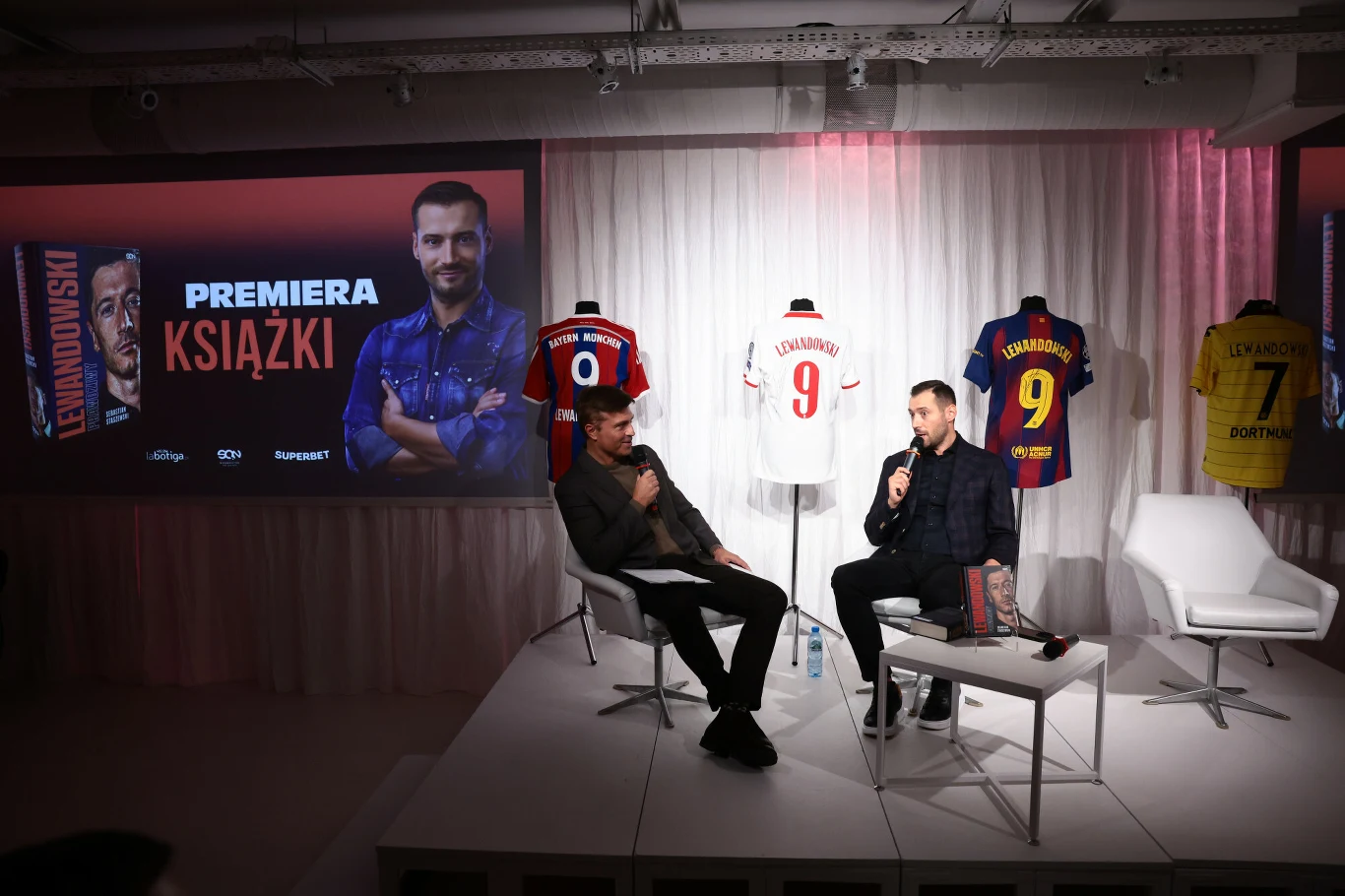 Dwóch mężczyzn prowadzi wywiad lub dyskusję na tle ekspozycji koszulek piłkarskich z nazwiskiem Lewandowski i numerami, obok wyświetlony plakat zapowiadający premierę książki