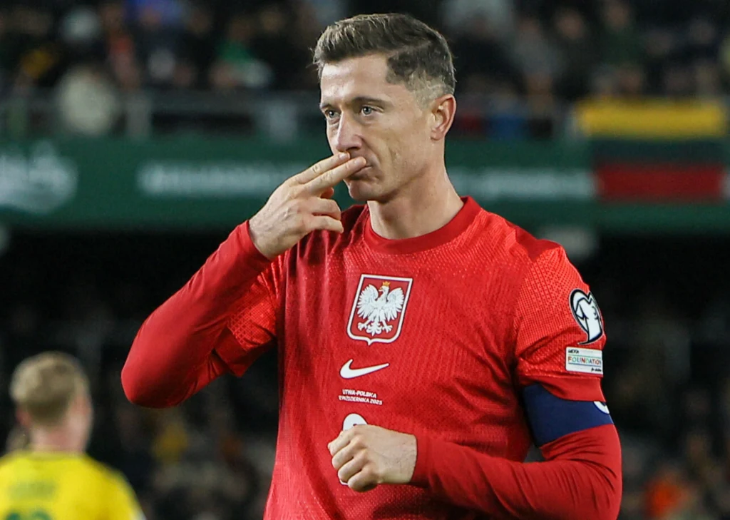 Robert Lewandowski w barwach reprezentacji Polski Piłkarz w czerwonej koszulce reprezentacji Polski wykonuje gest dłonią w kierunku ust. Na stroju widoczny jest herb Polski oraz opaska kapitana. W tle rozmyci zawodnicy i fragment stadionu.