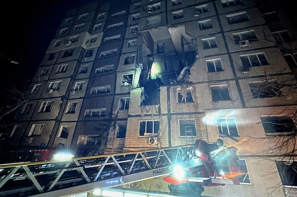 Nocna scena z uszkodzonym budynkiem wielorodzinnym, w którym jedna z kondygnacji została poważnie zniszczona, otwarte okna, spalone fragmenty elewacji; na pierwszym planie wysięgnik strażacki z ratownikami pracującymi przy miejscu zdarzenia.