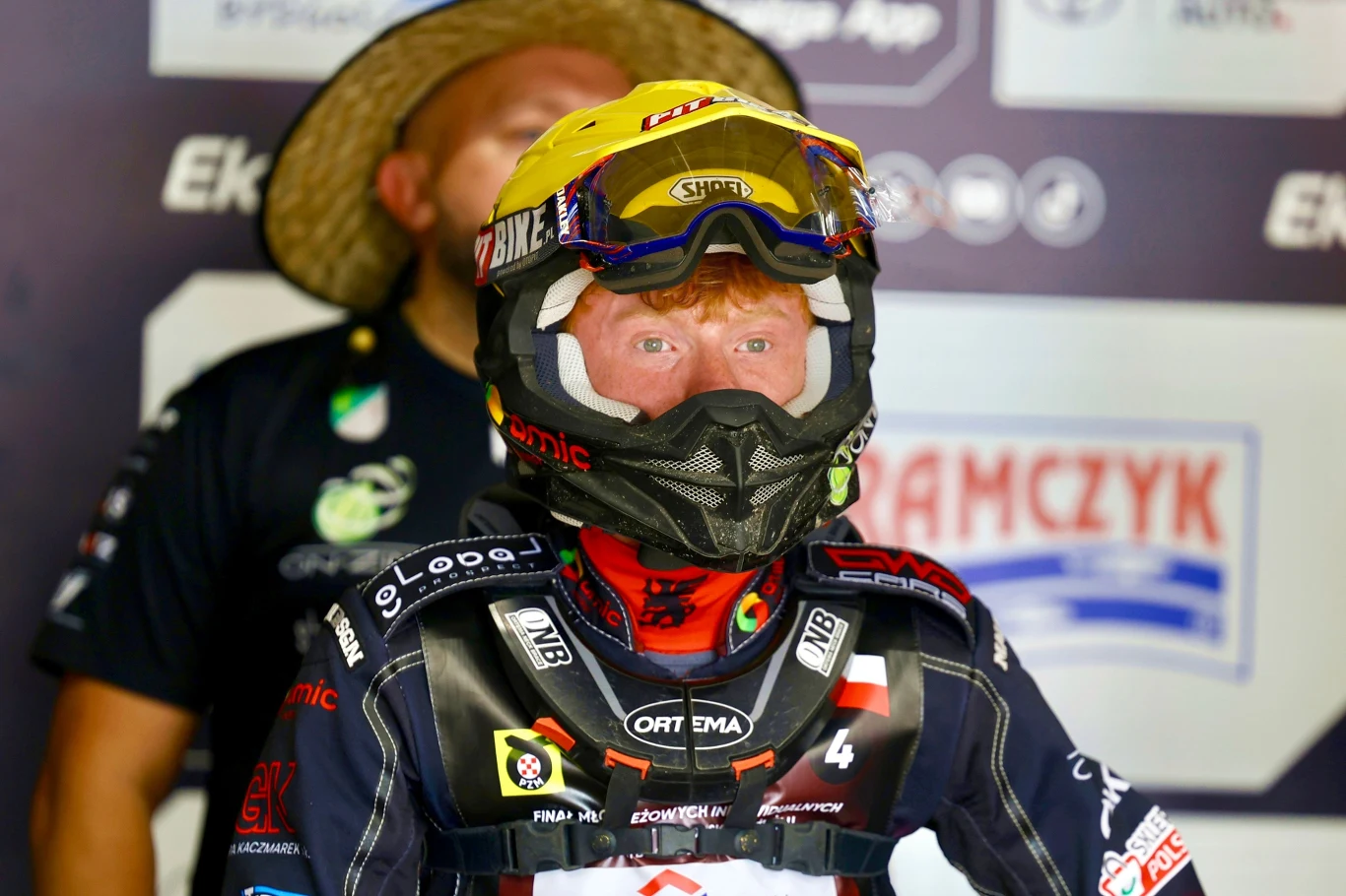 Maksymilian Pawełczak gotowy do startu. Mirosław Duda za jego plecami. Zawodnik żużlowy w kompletnym stroju ochronnym i kasku z goglami, stojący na tle tablicy z reklamami, w tle nieostro widoczny człowiek w kapeluszu.