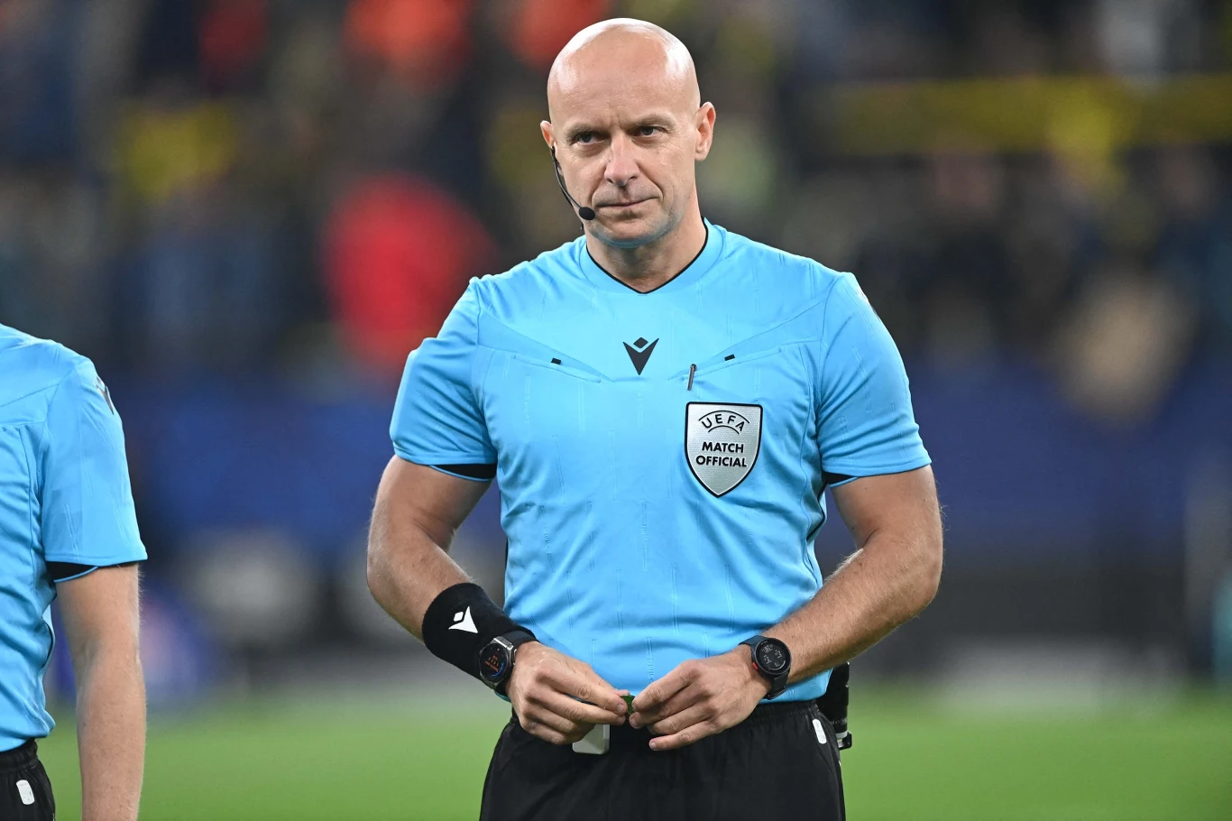 Szymon Marciniak Sędzia piłkarski w niebieskim stroju stoi na murawie boiska podczas meczu, w tle rozmyty widok trybun i kibiców.