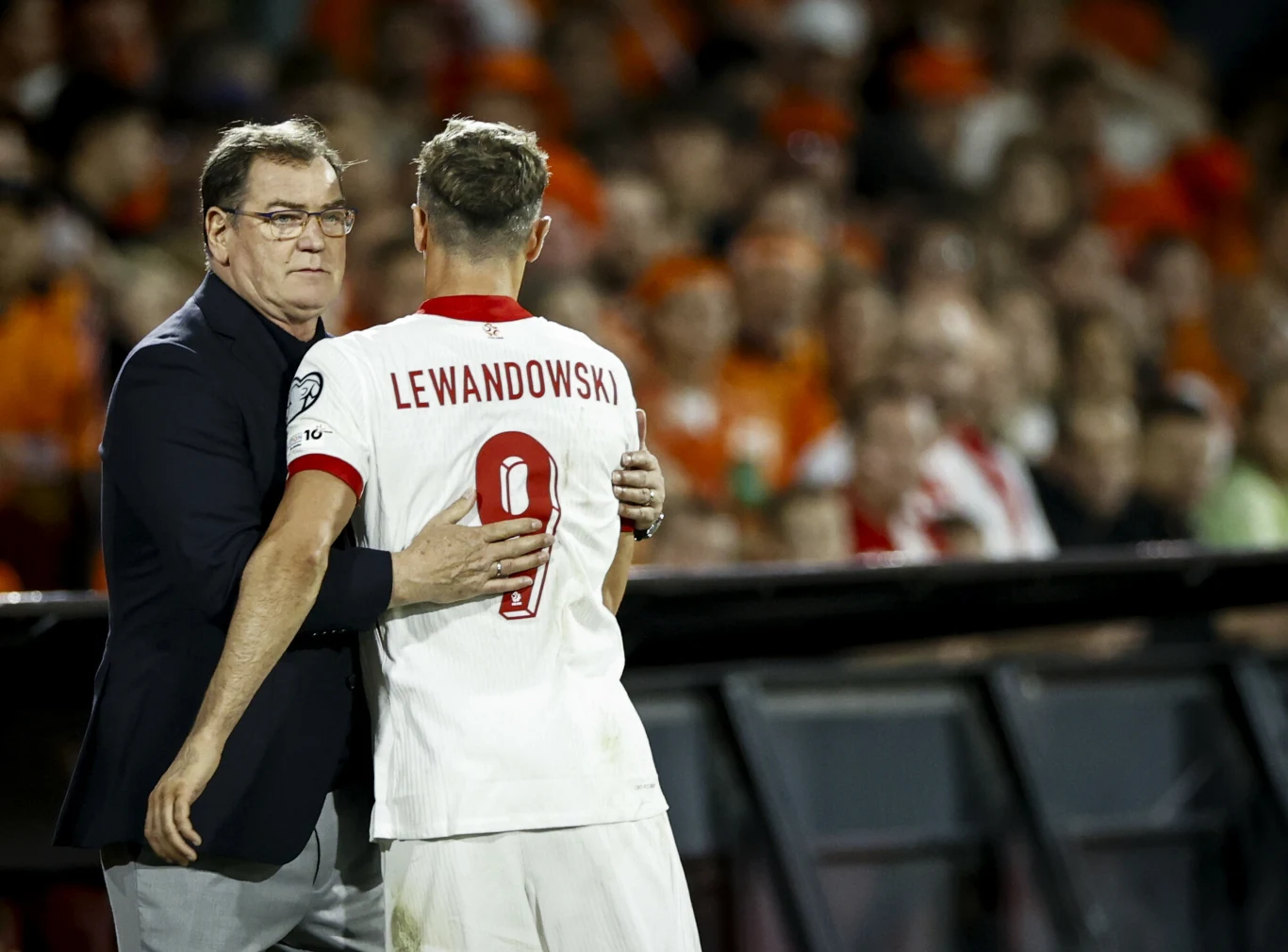 Trener w marynarce trzyma zawodnika w białej koszulce z nazwiskiem Lewandowski oraz numerem 9, w tle nieostry tłum kibiców w pomarańczowych barwach na stadionie piłkarskim.