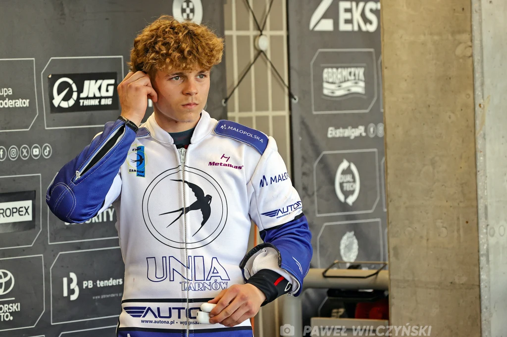 Radosław Kowalski w barwach Unii Tarnów. Młody mężczyzna w stroju żużlowca z logo Unii Tarnów, przygotowujący się do zawodów, na tle ściany z logotypami sponsorów.