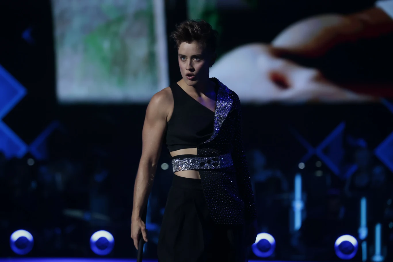 Osoba w wyrazistym stroju scenicznym z jednym ramiączkiem i połyskującym elementem stoi na scenie na tle ekranu z rozmazanym obrazem. Oświetlenie skupia się na postaci, podkreślając dramatyzm występu.