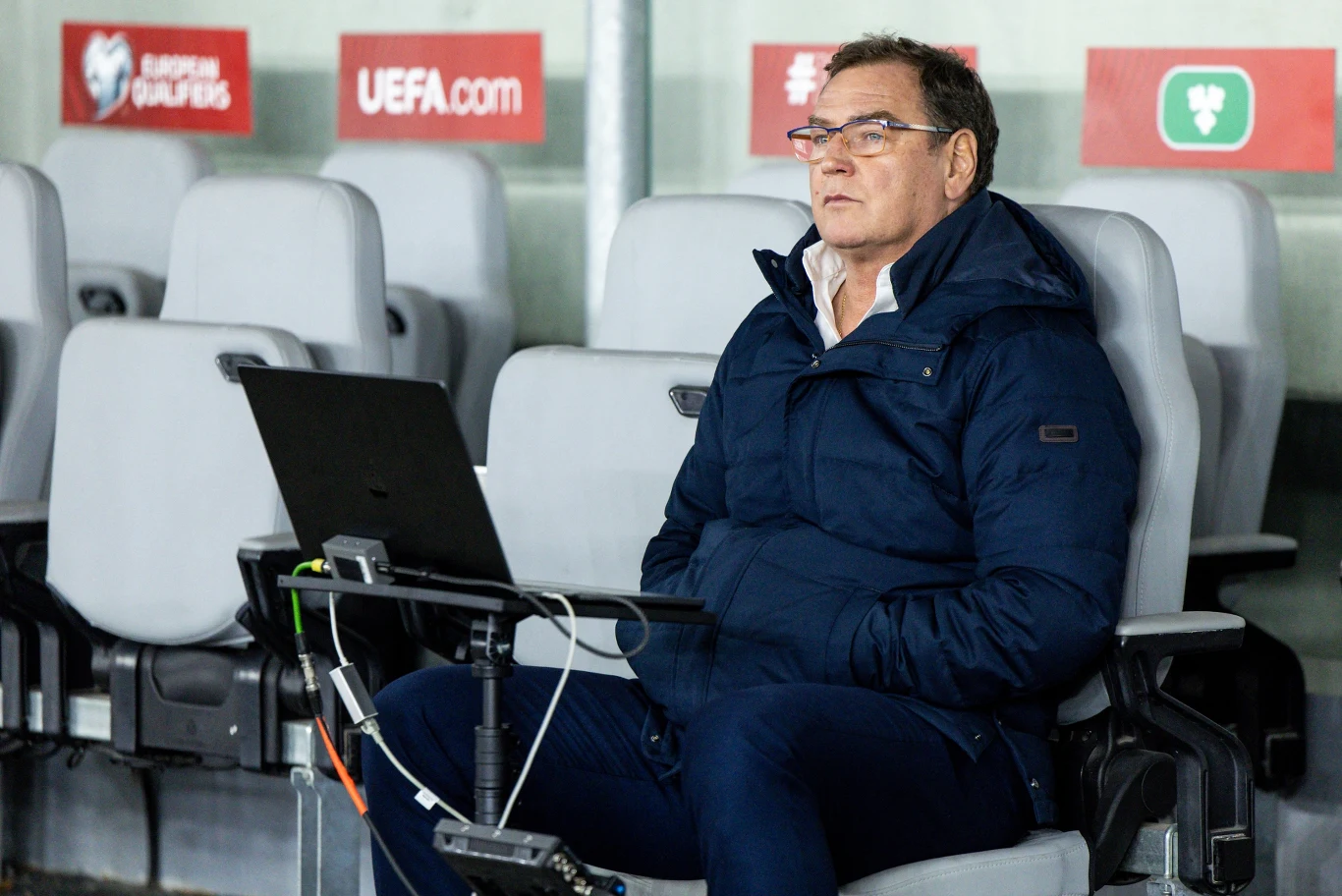 Jan Urban Mężczyzna w okularach siedzi samotnie na stadionowych krzesełkach, ubrany w granatową kurtkę i ciemne spodnie, przed nim znajduje się otwarty laptop na stoliku, w tle widoczne są logotypy UEFA.