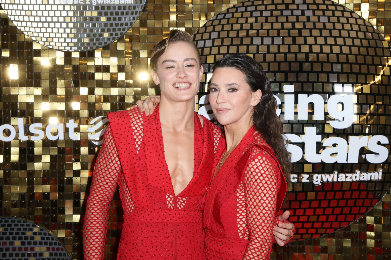 Dwie kobiety w efektownych, czerwonych sukniach stoją blisko siebie i pozują do zdjęcia na tle złotej ścianki z logo programu telewizyjnego Dancing with the Stars.