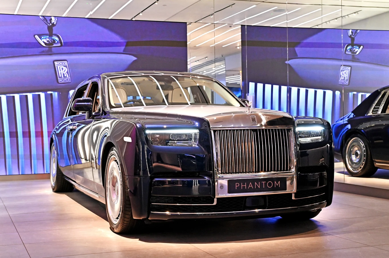 Luksusowy samochód Rolls-Royce Phantom w ciemnogranatowym kolorze stoi w nowoczesnym salonie na tle wielkoformatowych ekranów prezentujących logo producenta i detale pojazdu.