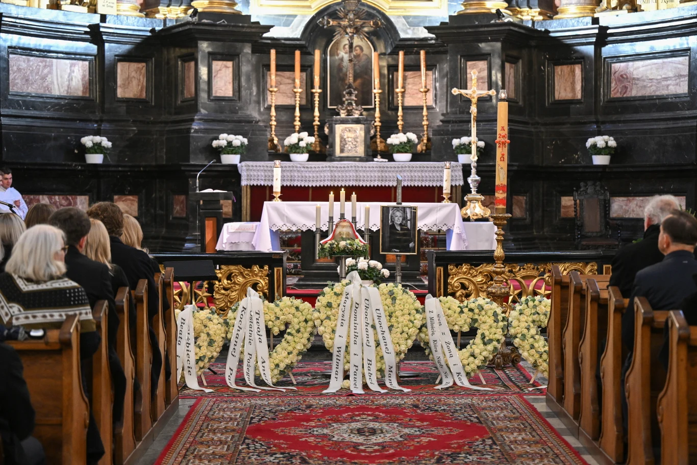 Wnętrze kościoła podczas uroczystości pogrzebowej, w centralnej części ołtarz otoczony licznymi wieńcami i kwiatami, zdjęcie zmarłego oraz szarfy żałobne, zebrani ludzie siedzą w ławkach zwróceni w stronę ołtarza.