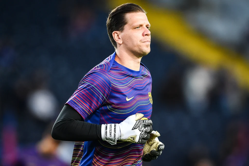 Wojciech Szczęsny Mężczyzna w sportowej koszulce piłkarskiej i rękawicach bramkarskich podczas rozgrzewki na stadionie, w tle rozmyte trybuny.