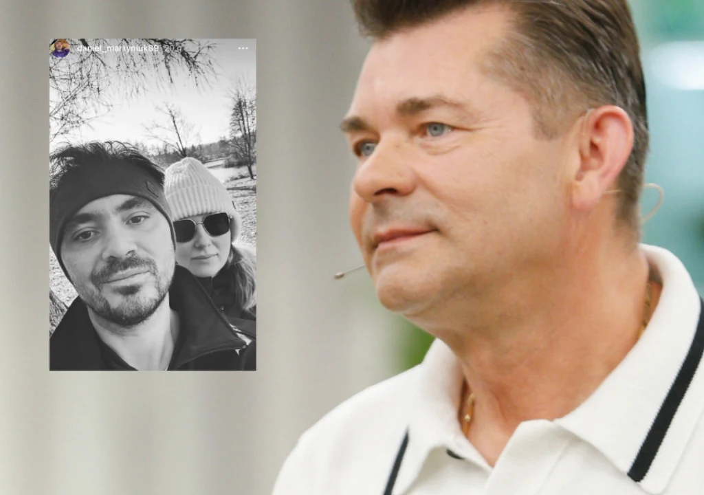 Zbliżenie mężczyzny w białej koszuli z mikrofonem na uchu, w tle widoczna czarno-biała fotografia przedstawiająca dwie osoby, mężczyznę i kobietę w czapkach zimowych.