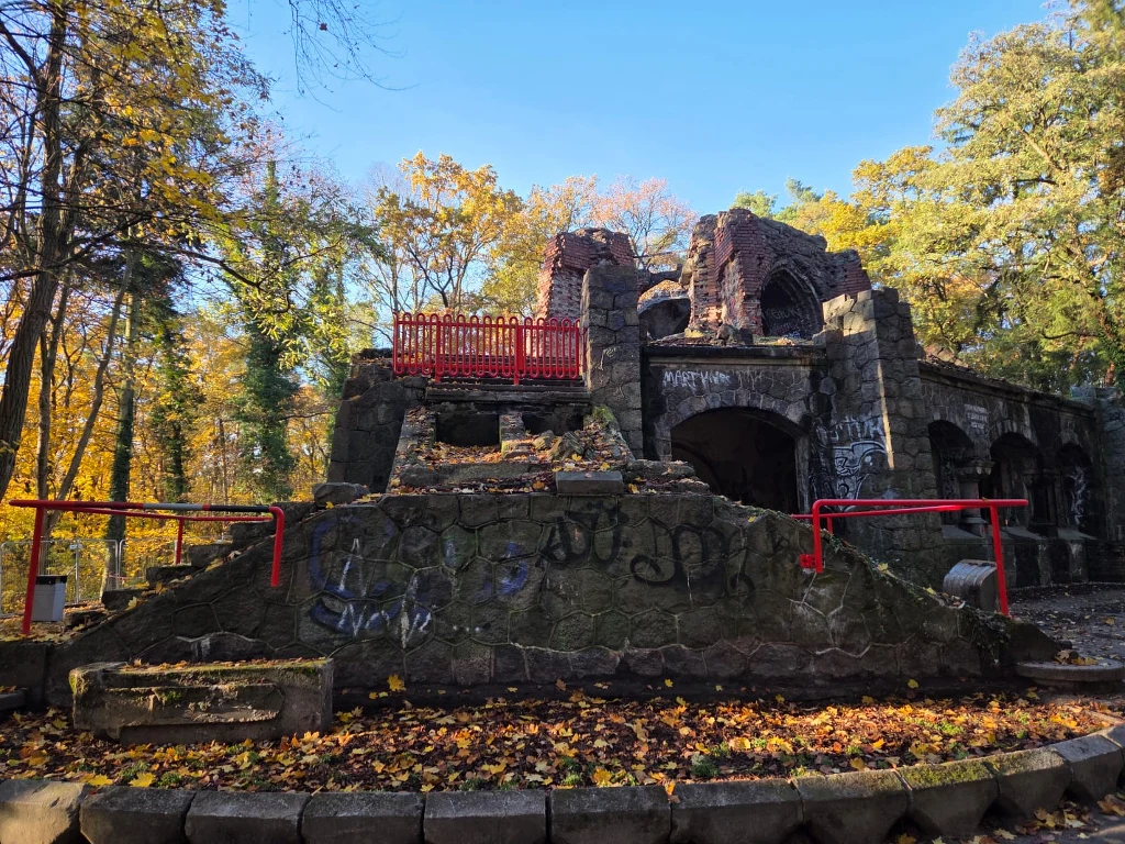 Ruiny kamiennego budynku otoczone jesiennym lasem, na ścianach widoczne graffiti, kilka metalowych poręczy w kolorze czerwonym kontrastuje z szarością konstrukcji, całość otoczona liśćmi i naturalną roślinnością.