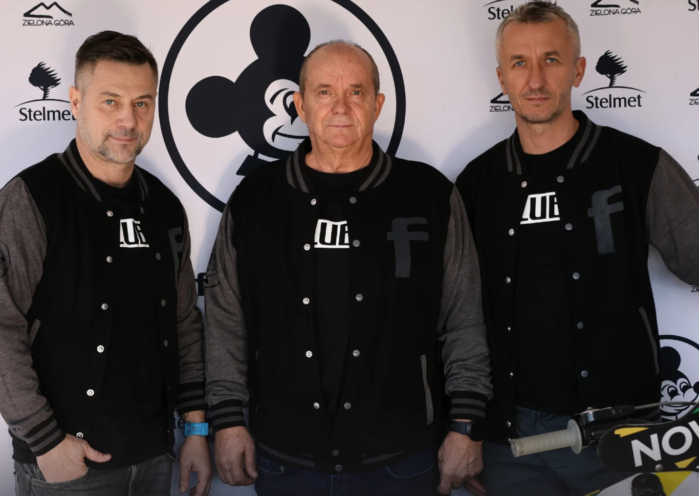 Prezes Falubazu Adam Goliński, dyrektor sportowy Aleksander Janas oraz menedżer Grzegorz Walasek Trzech mężczyzn w czarnych kurtkach z logo, stojących przed tłem z symbolem myszy oraz logotypami sponsorów. Jeden z mężczyzn opiera rękę na rowerze, całość utrzymana w neutralnym tonie.