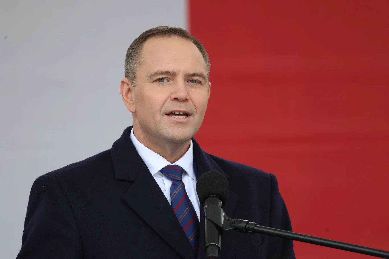 Mężczyzna w garniturze przemawia do mikrofonu na tle biało-czerwonej flagi, prezentując poważny wyraz twarzy.