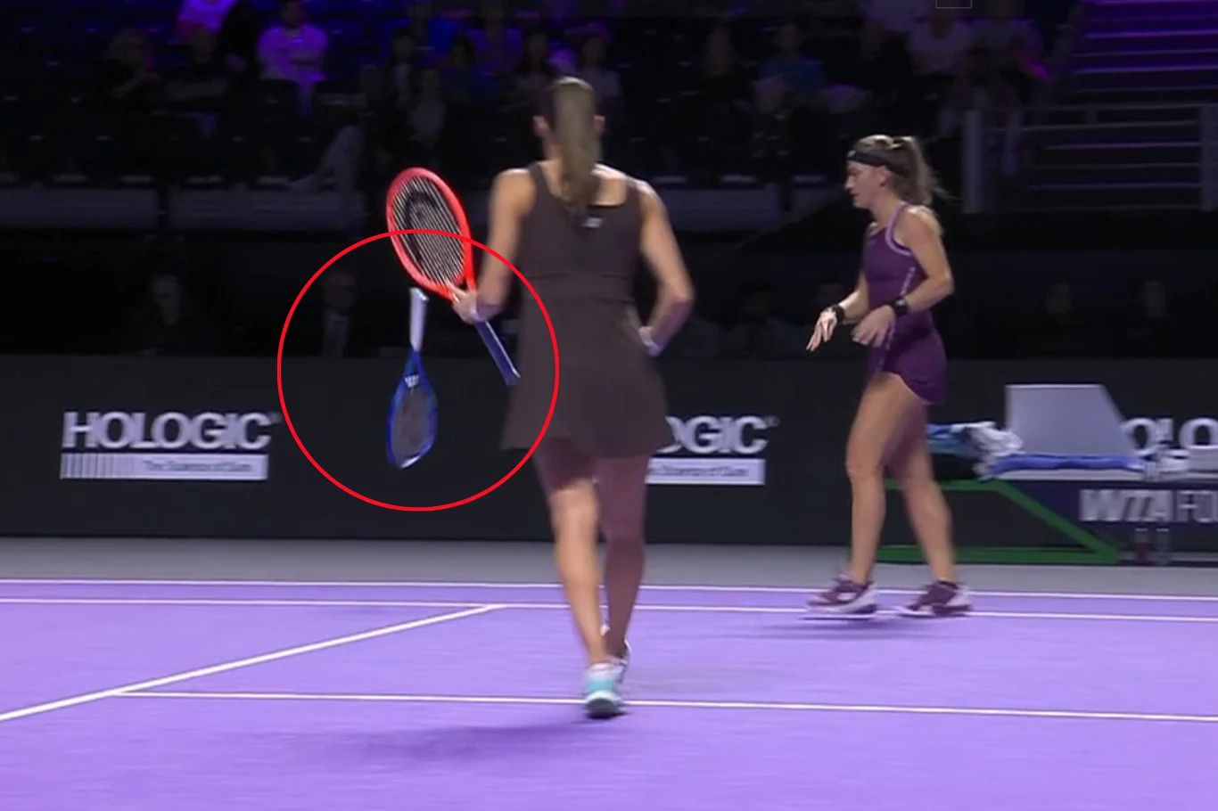 Zawodniczka na korcie tenisowym spuszczająca rakietę, w tle druga tenisistka oraz ławka na bocznej linii. Kort ma fioletową nawierzchnię, a na bandach widoczne są logotypy sponsorów.