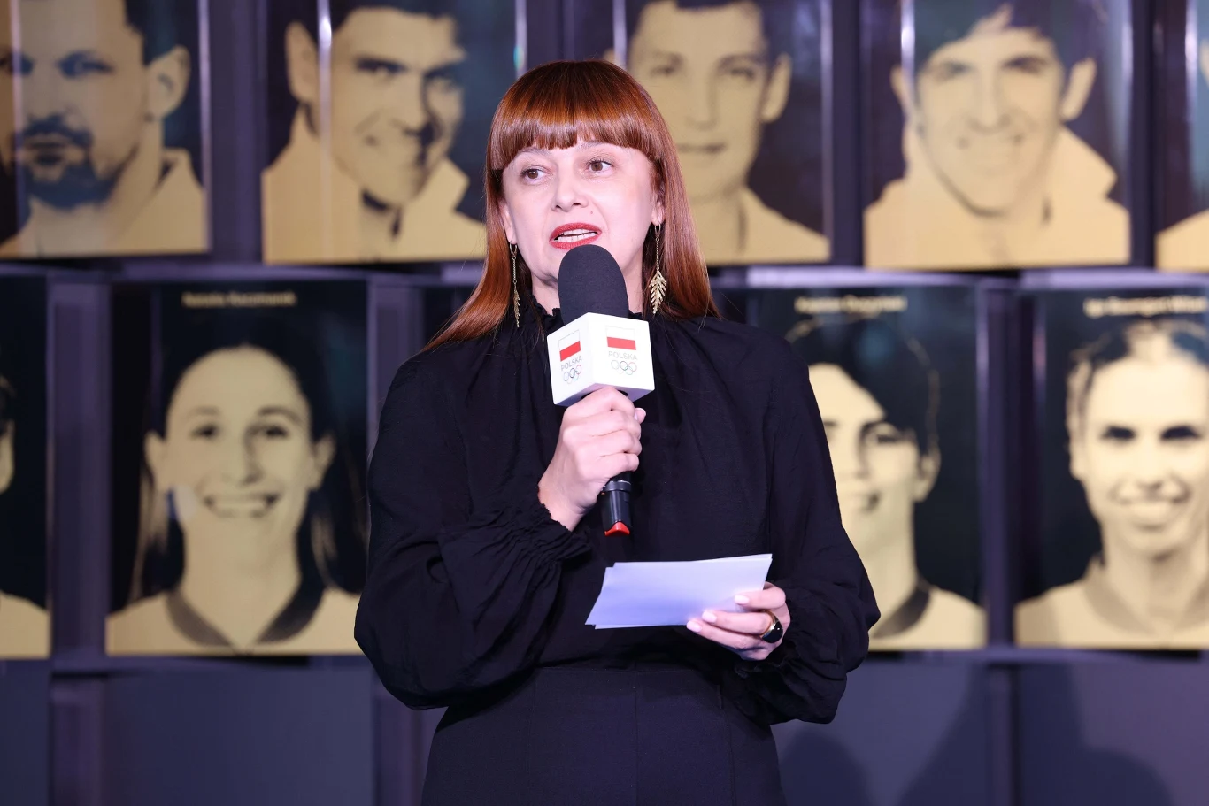 Hanna Wawrowska Kobieta z rudymi włosami w czarnej sukience przemawia do mikrofonu, trzymając kartkę, w tle widoczne portrety kilku osób na żółtych planszach.