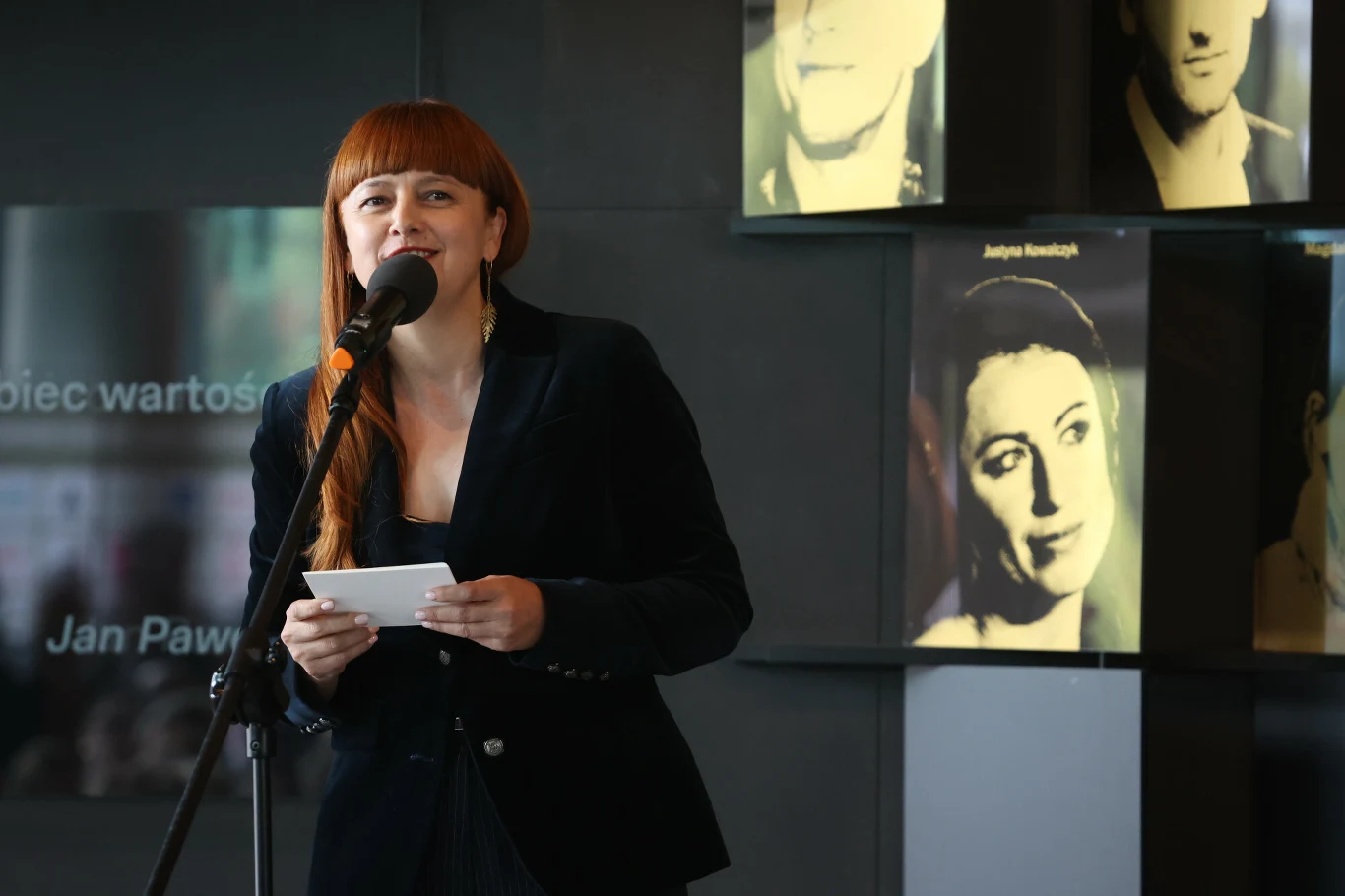 Hanna Wawrowska Kobieta z rudymi włosami stoi przy mikrofonie, trzymając kartkę papieru. Za nią znajdują się zdjęcia portretowe znanych osób, umieszczone na ścianie.