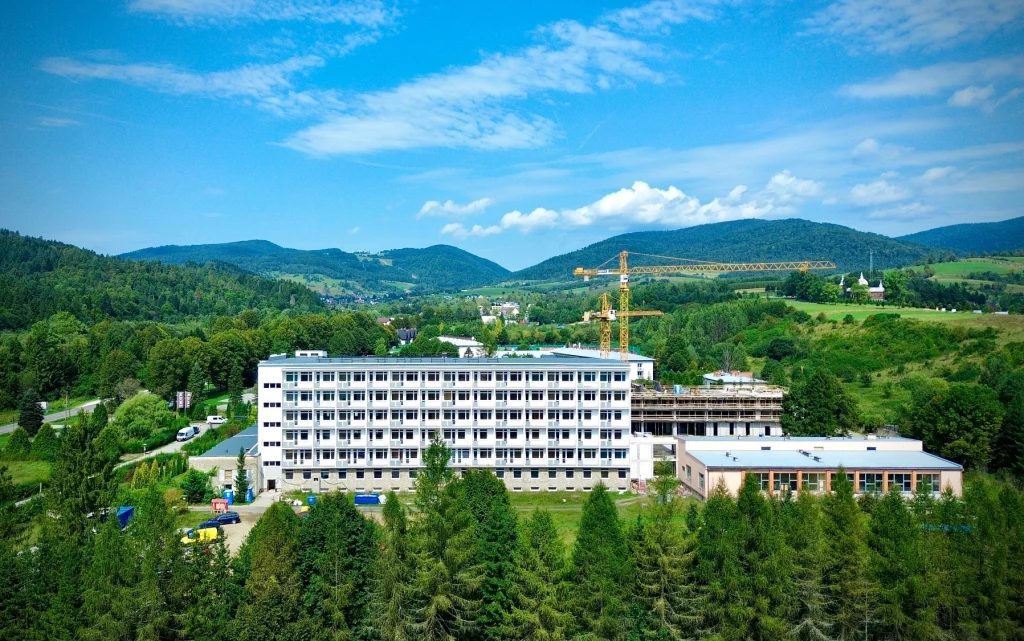 Budynek wielopiętrowy otoczony zieloną roślinnością i drzewami, w tle góry oraz fragment żółtego żurawia budowlanego wskazujący na prowadzone prace budowlane.