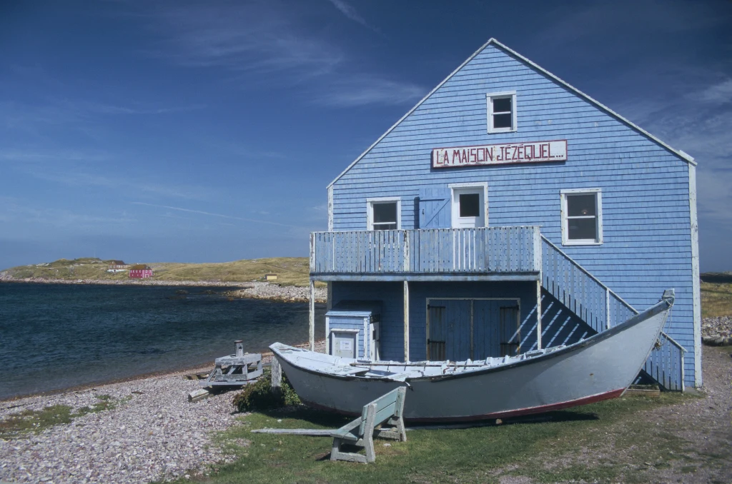 W wiosce Miquelon nie można już stawiać domów zbyt blisko poziomu morza Niebieski drewniany dom z tablicą La Maison Jezebel, zlokalizowany nad brzegiem zbiornika wodnego, z dużą łodzią ustawioną przed wejściem oraz ławkami na kamienistym podwórzu, w tle rozciąga się krajobraz nadmorski.