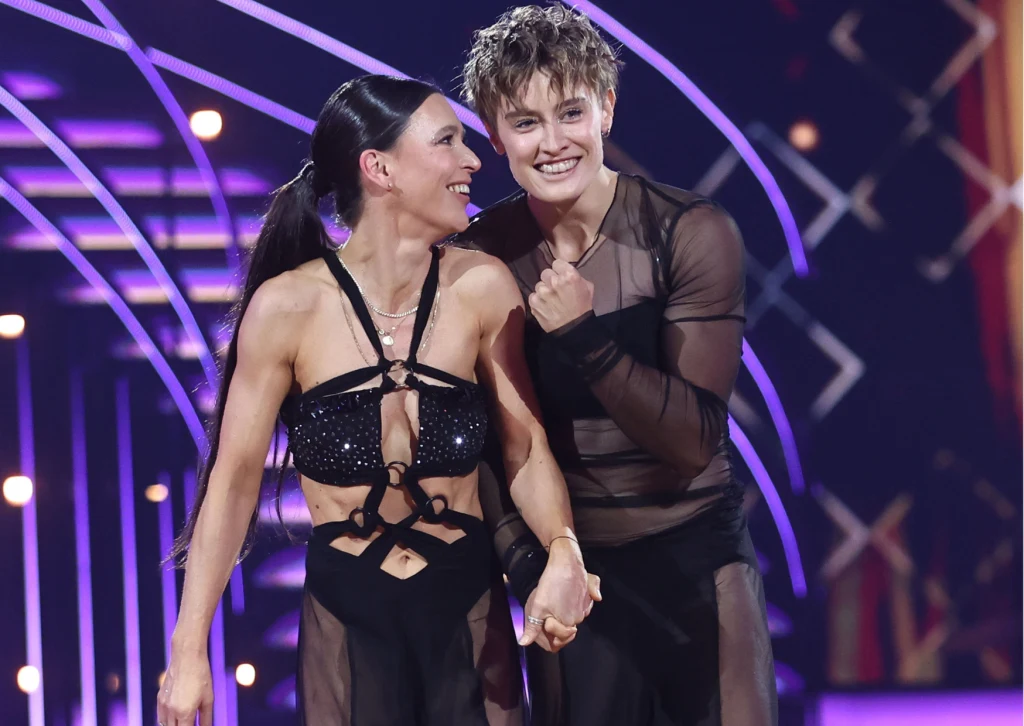 Dwoje uśmiechniętych tancerzy trzyma się za ręce na scenie oświetlonej kolorowymi światłami, oboje ubrani są w efektowne, czarne stroje sceniczne z przezroczystymi elementami.