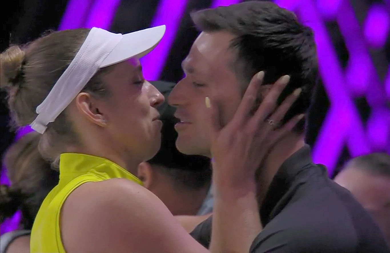 Dwie osoby zwracają się ku sobie, trzymając się za twarze z zamkniętymi oczami, jakby za chwilę miały się pocałować. Kobieta w białej czapce z daszkiem i żółtej koszulce dotyka policzka mężczyzny. W tle widoczne są fioletowe światła oraz niewyraźne pos...