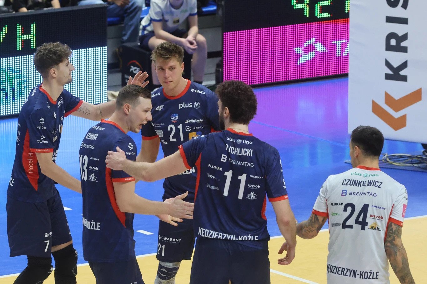 Grupa siatkarzy z drużyny Grupa Azoty ZAKSA Kędzierzyn-Koźle świętuje zdobycie punktu na parkiecie hali sportowej, zawodnicy są ubrani w klubowe stroje, wokół nich widoczna część boiska oraz fragment elektronicznej tablicy wyników.