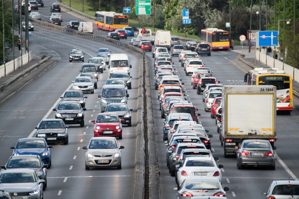 repo Zatłoczona miejska arteria z natężonym ruchem samochodów w obu kierunkach, widoczne samochody osobowe i ciężarówka, w tle jadące autobusy komunikacji miejskiej oraz miejskie oznakowanie drogowe.
