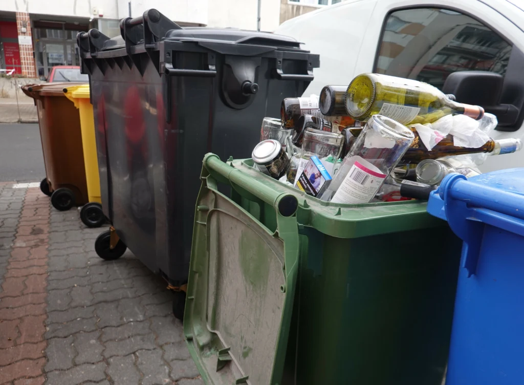 Niektóre gminy w Polsce wymagają, aby szkło wyrzucać do śmieci bez worków foliowych Przepełniony pojemnik na szkło z dużą ilością pustych butelek i słoików, ustawiony obok innych pojemników na odpady na chodniku w pobliżu budynku mieszkalnego.