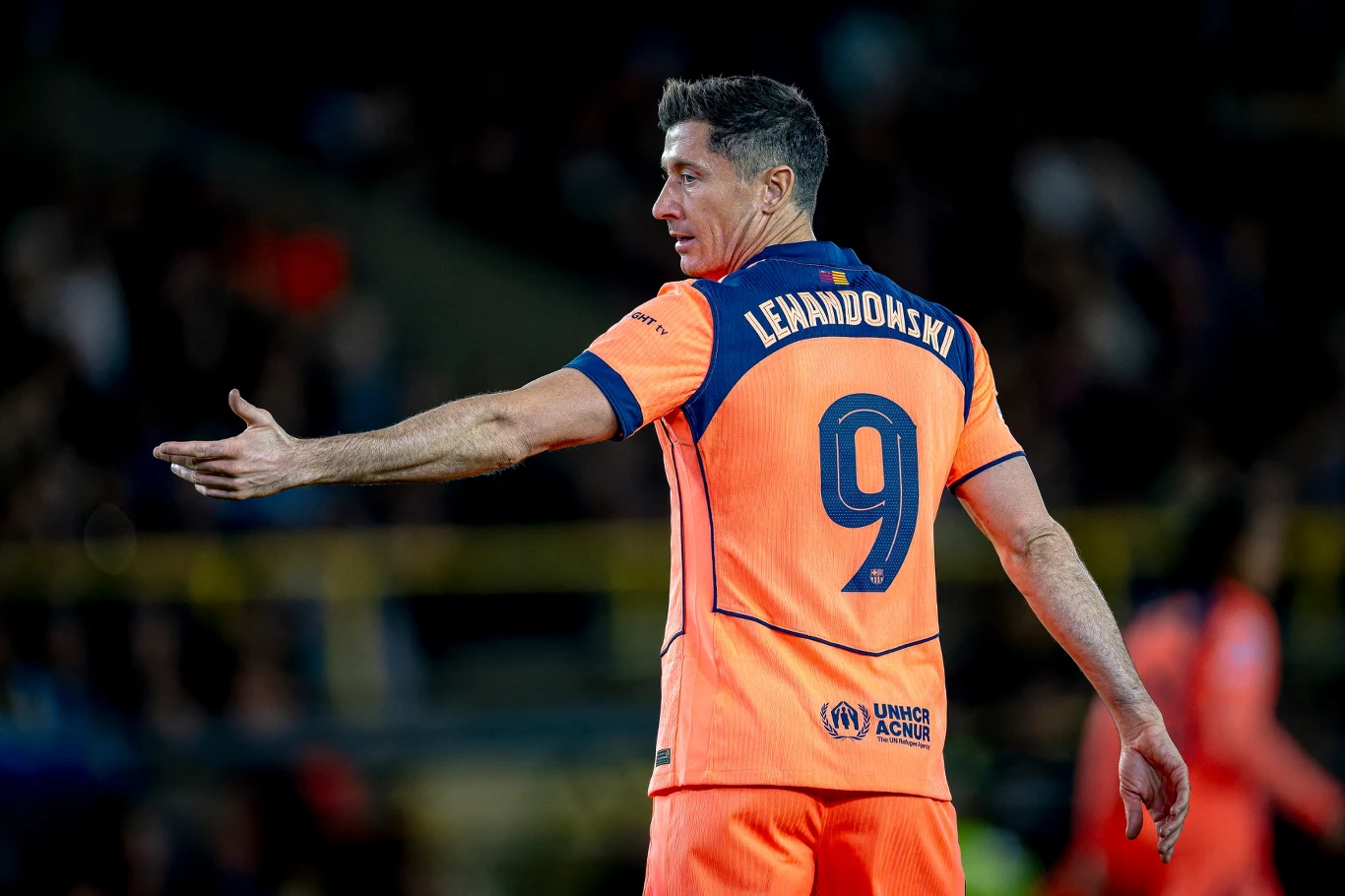 Piłkarz w pomarańczowym stroju sportowym z numerem 9 i nazwiskiem Lewandowski na plecach, gestykuluje ręką podczas meczu piłkarskiego, widoczny rozmyty tłum w tle.