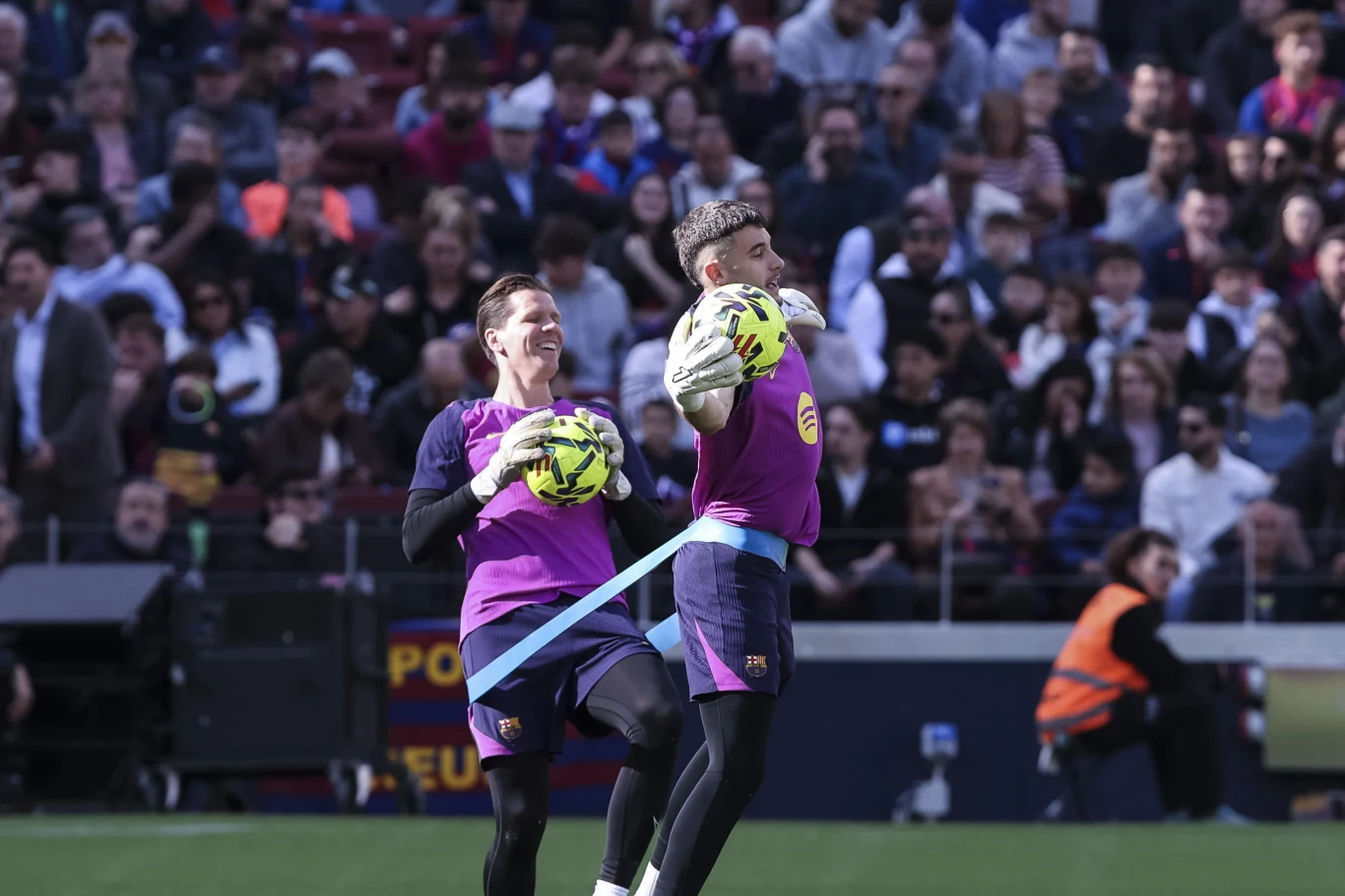Dwóch bramkarzy drużyny piłkarskiej podczas treningu na stadionie, połączeni niebieską taśmą, każdy z piłką w rękach, wokół liczna publiczność na trybunach.