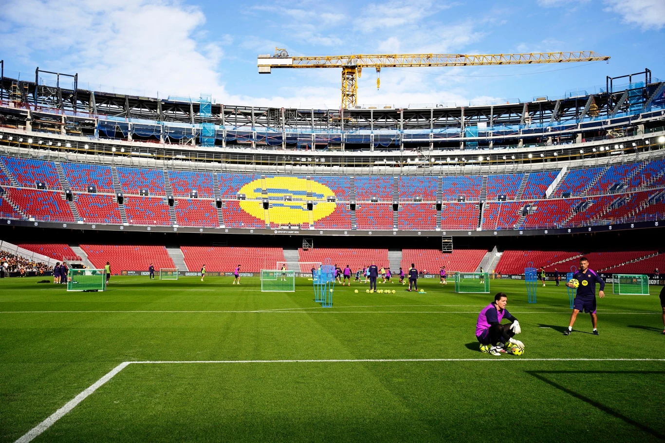 Camp Nou Stadion piłkarski wypełniony kolorowymi krzesełkami, z widocznym żółtym kształtem na trybunach, zawodnicy trenujący na murawie, w tle żuraw budowlany nad górą stadionu, puste trybuny.