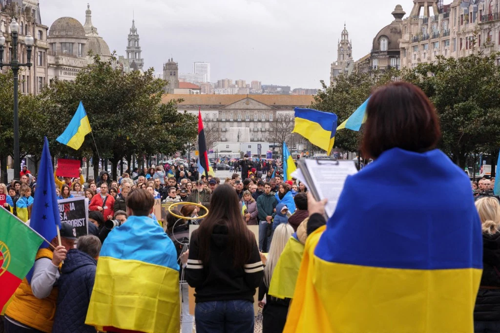 Protest społeczności ukraińskiej w Porto, luty 2024 roku. Uchodźcy z Ukrainy mogą stracić swój status - donoszą portugalskie media Tłum ludzi zgromadzonych na miejskim placu podczas demonstracji, widoczne są liczne flagi Ukrainy oraz transparenty, jedna z osób ma flagę owiniętą na plecach i odczytuje coś do zgromadzenia, w tle architektura miejskich kamienic.