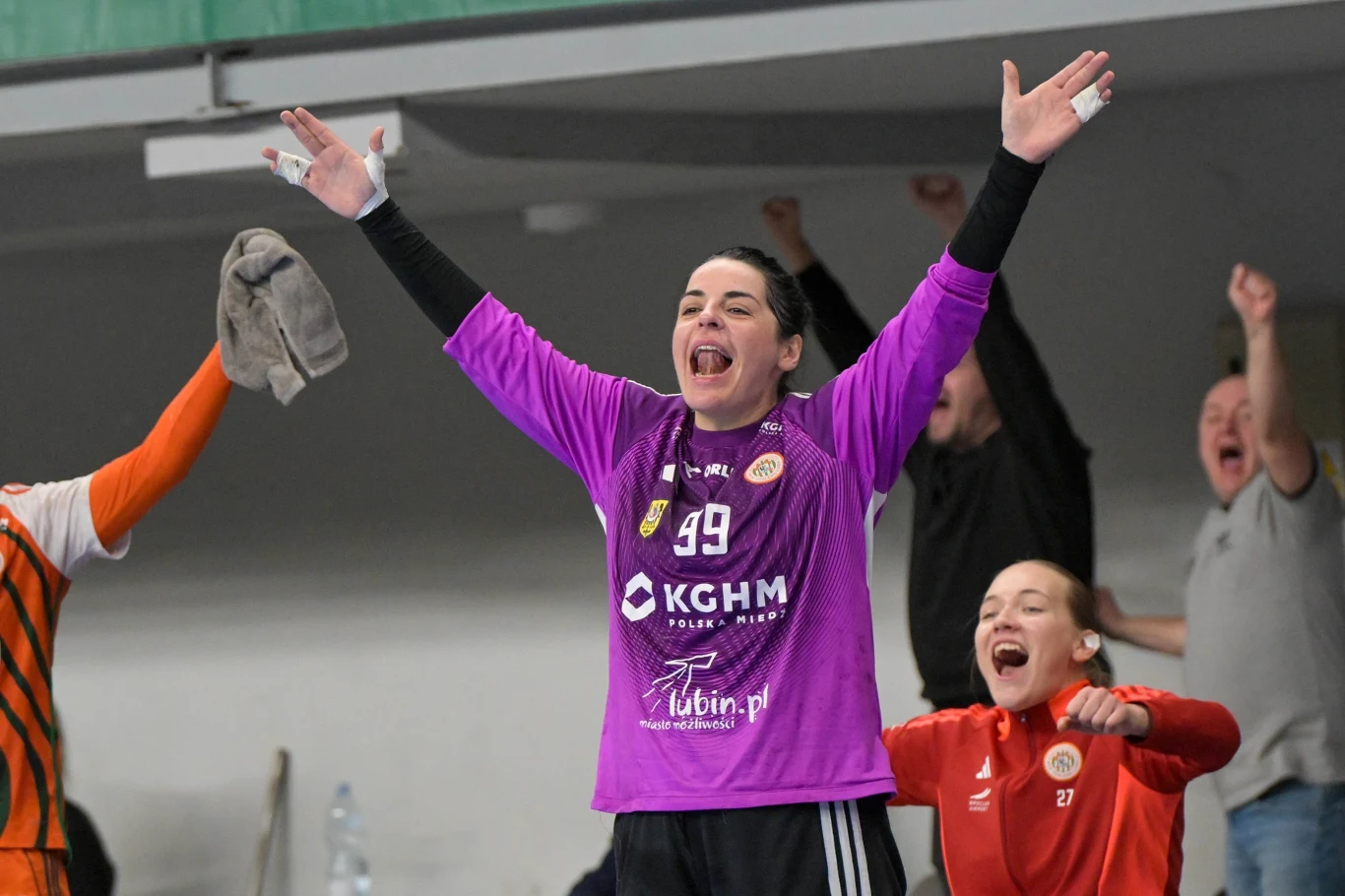 Zawodniczka w fioletowej koszulce z szeroko uniesionymi ramionami celebruje sukces wraz z innymi osobami w tle, wszyscy wyrażają radość i entuzjazm po udanym wydarzeniu sportowym.