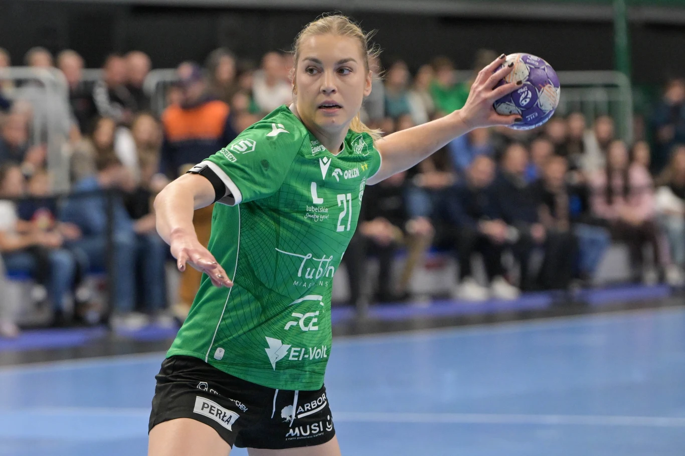 Zawodniczka piłki ręcznej w zielonej koszulce przygotowuje się do rzutu podczas meczu, w tle widoczna publiczność na trybunach.