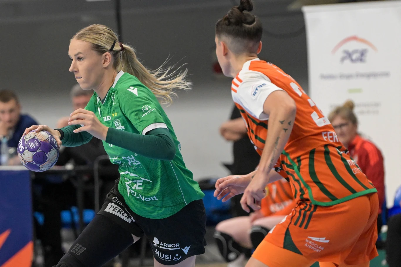 Dwie zawodniczki rywalizujące podczas meczu piłki ręcznej, jedna ubrana w zieloną, druga w pomarańczową koszulkę, obie skupione na piłce i akcji. W tle sędziowie oraz publiczność.