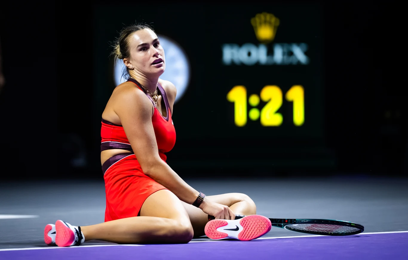 Tenisistka siedzi na korcie w sportowej czerwonej sukience z rakietą leżącą obok niej, w tle duży zegar oraz marka Rolex.