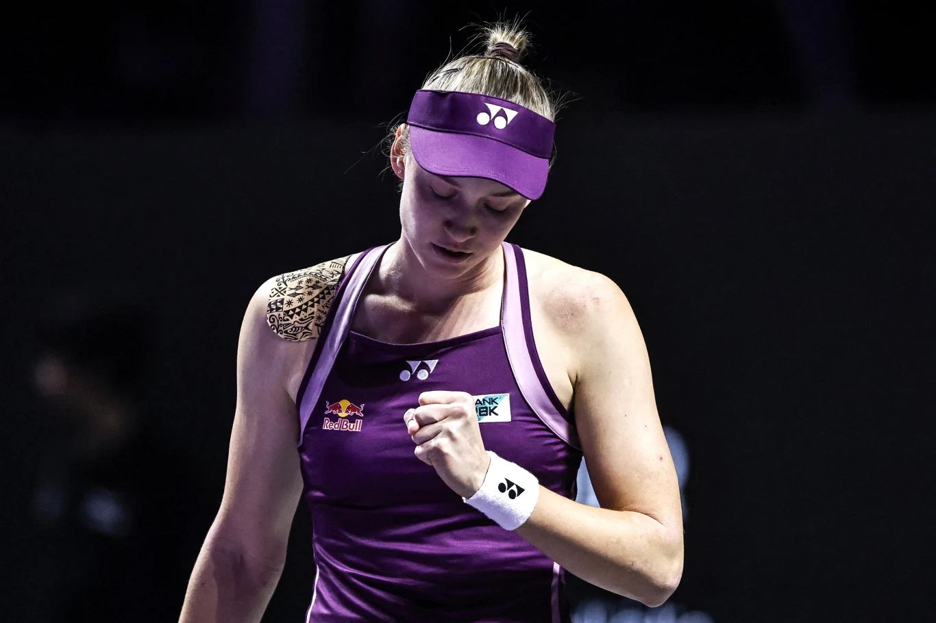 Zawodowa tenisistka w fioletowym stroju sportowym z opaską na głowie i plastrem na prawym ramieniu zaciska pięść w geście determinacji podczas meczu tenisowego.