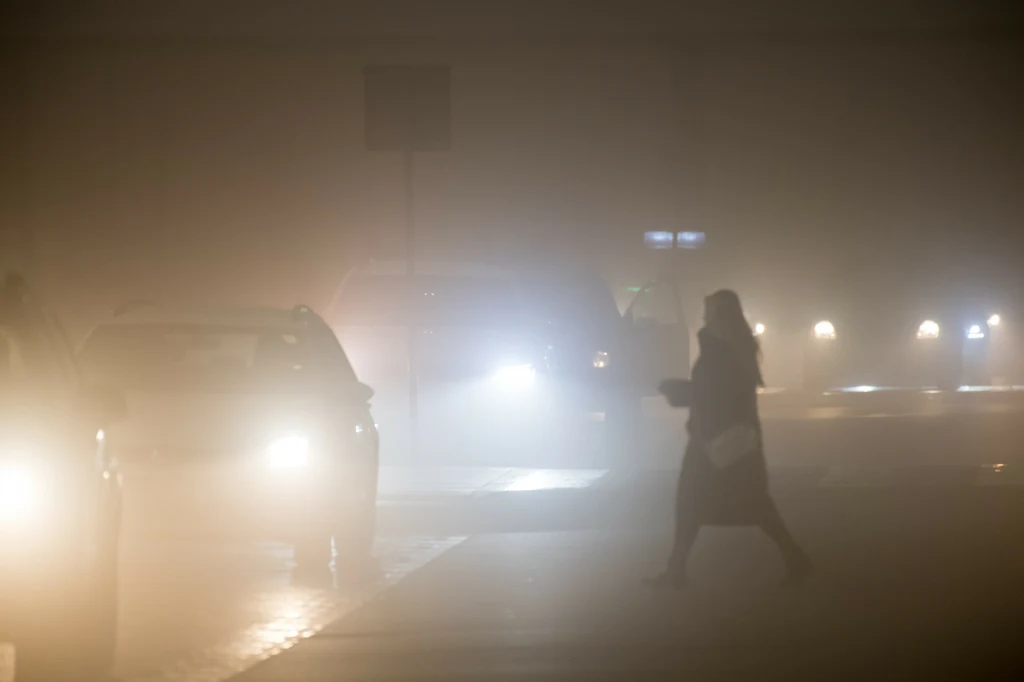 Gęsta mgła znacznie ograniczy widoczność. IMGW wydał ostrzeżenia. Zdj. ilustracyjne Silnie zacieniona postać człowieka przechodząca przez jezdnię w gęstej mgle, zbliżające się samochody z włączonymi światłami, ograniczona widoczność na drodze.