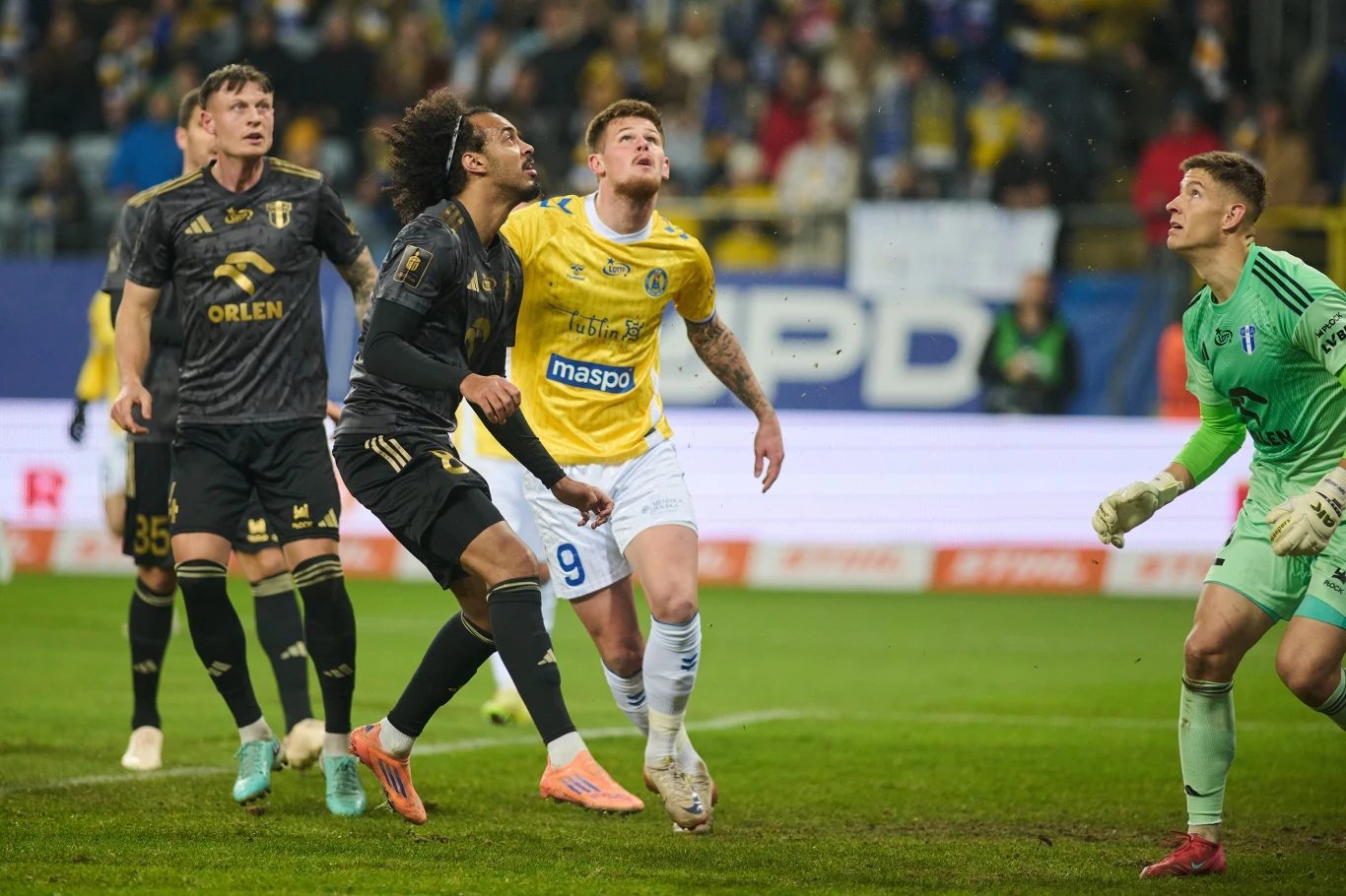 Motor Lublin - Wisła Płock Piłkarze dwóch drużyn skupieni wokół bramki w trakcie dynamicznej akcji podczas meczu piłki nożnej, widoczny bramkarz przygotowany do obrony, zawodnicy w żółtych i czarnych strojach rywalizują o piłkę, na trybunach tłum kibiców.