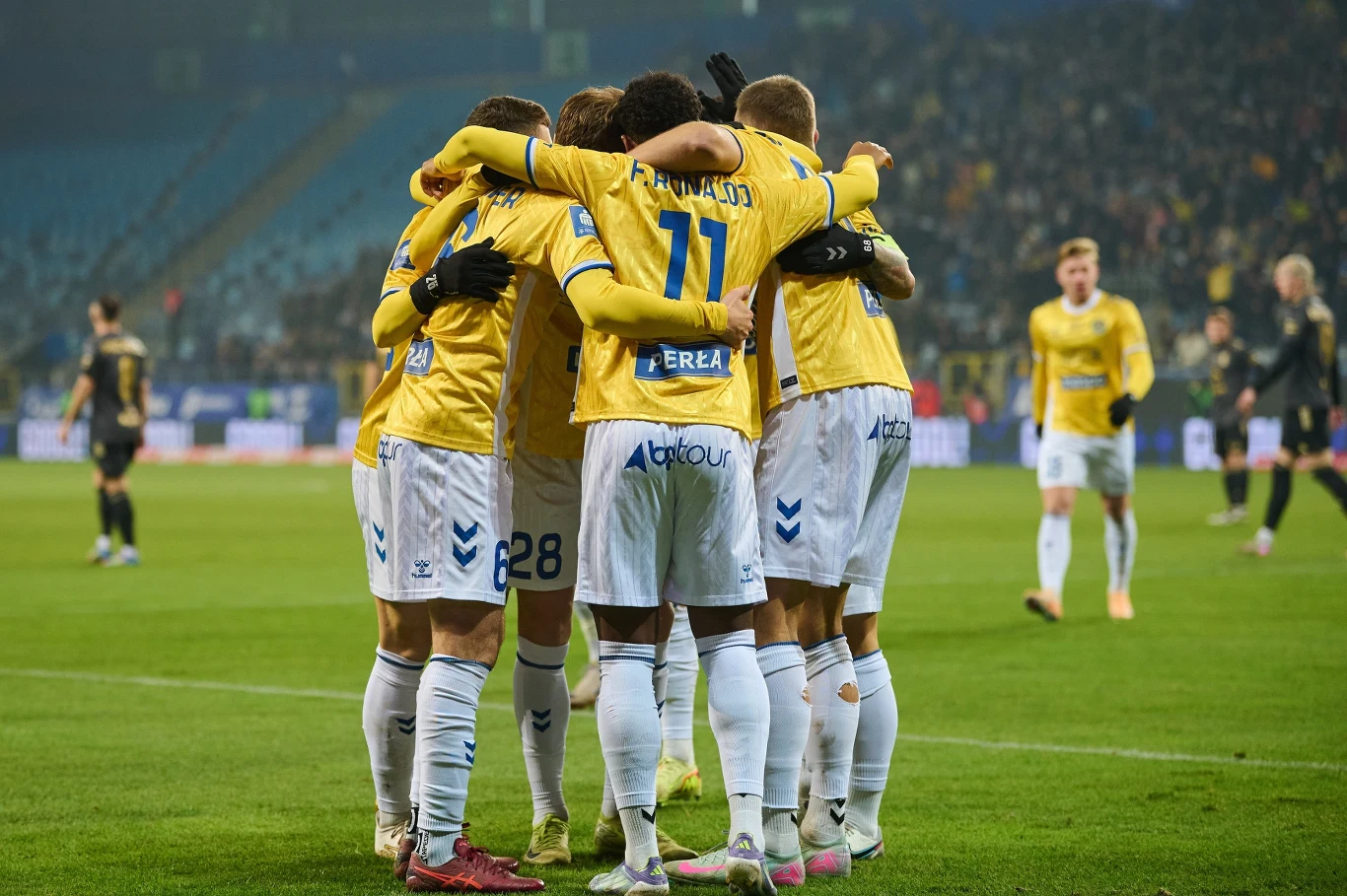 Motor Lublin Grupa piłkarzy w żółtych koszulkach i białych spodenkach świętuje zdobycie gola, przytulając się na środku boiska, w tle widać rozmytą publiczność oraz innych zawodników.