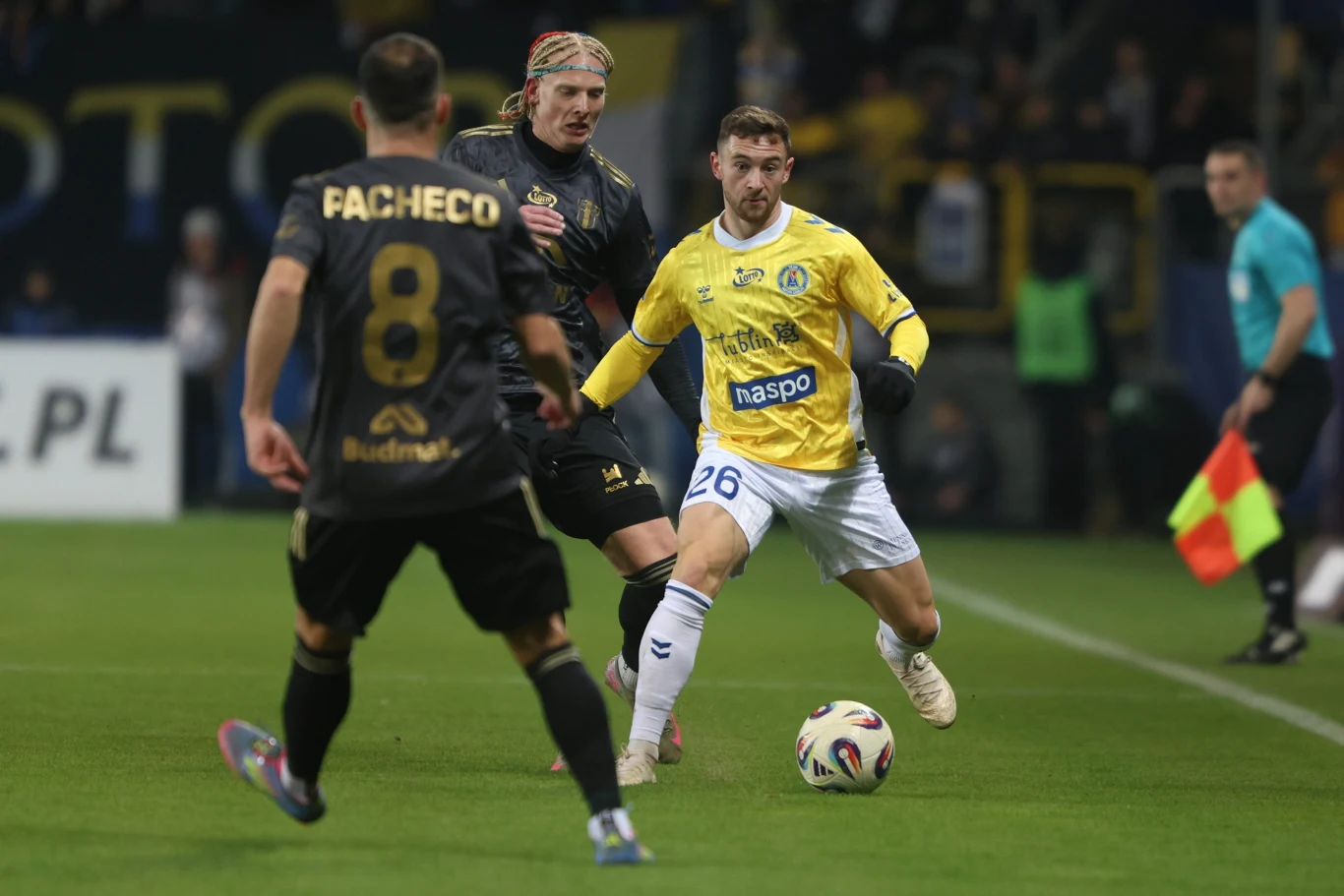 Motor Lublin Zawodnik w żółtej koszulce prowadzi piłkę drużyny piłkarskiej, próbując minąć dwóch przeciwników ubranych w czarne stroje, w tle widać liniowego sędziego z chorągiewką na murawie oświetlonego stadionu.