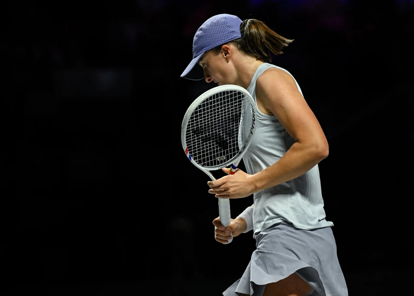 Tenisistka w sportowym stroju z rakietą tenisową w dłoni na tle ciemnego boiska, skoncentrowana i w ruchu, w sportowej czapce i jasnej koszulce.