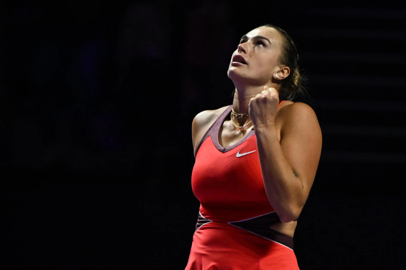 Aryna Sabalenka Tenisistka w czerwonej sukience wykonująca gest triumfu ze zaciśniętą pięścią, jej twarz wyraża emocje i skupienie, tło zaciemnione bez widocznych szczegółów.