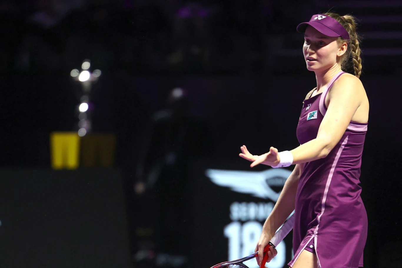 Tenisistka ubrana w fioletowy strój sportowy gestykuluje podczas meczu na korcie, na drugim planie widoczny zamazany puchar.