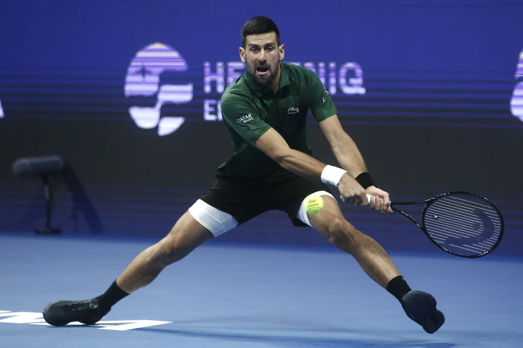 Novak Djoković Tenisista wykonuje szeroki rozkrok podczas gry na niebieskim korcie, trzymając rakietę tuż przy piłce tenisowej, z wyrazem koncentracji na twarzy. W tle widoczne logo turnieju.