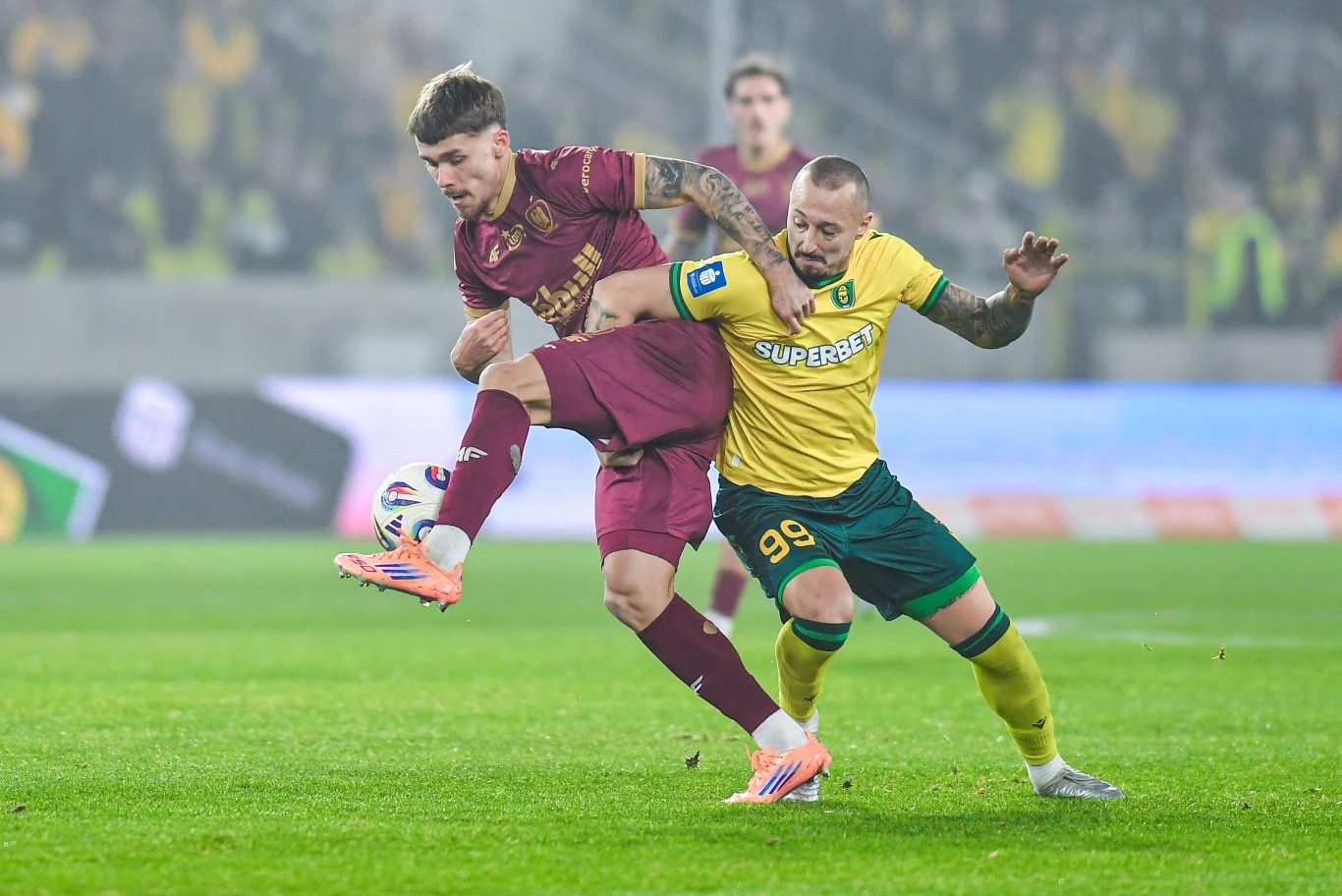 Dwóch piłkarzy rywalizuje o piłkę na boisku podczas meczu, jeden ubrany w bordowy strój, drugi w żółto-zielony trykot. Intensywna walka o posiadanie piłki odbywa się na tle rozmytej publiczności i stadionowego oświetlenia.