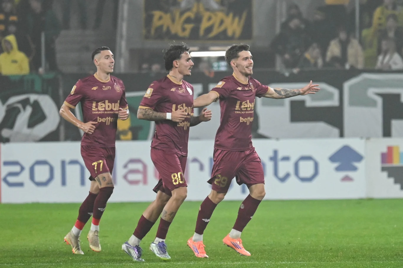 Trzech piłkarzy w bordowych strojach z numerami 77, 80 i 29 biegnie po murawie, dwóch z nich uśmiecha się i wykonuje gest radości po zdobyciu gola, w tle widoczny rozmyty tłum kibiców oraz fragment transparentu.