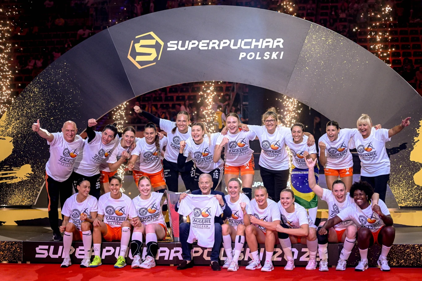 Radosna drużyna piłkarek oraz ich sztab trenerski celebrują zwycięstwo na podwyższeniu pod łukiem z napisem Superpuchar Polski. Wszyscy ubrani w białe koszulki triumfu, wokół dekoracje ze sztucznymi ogniami i medale.